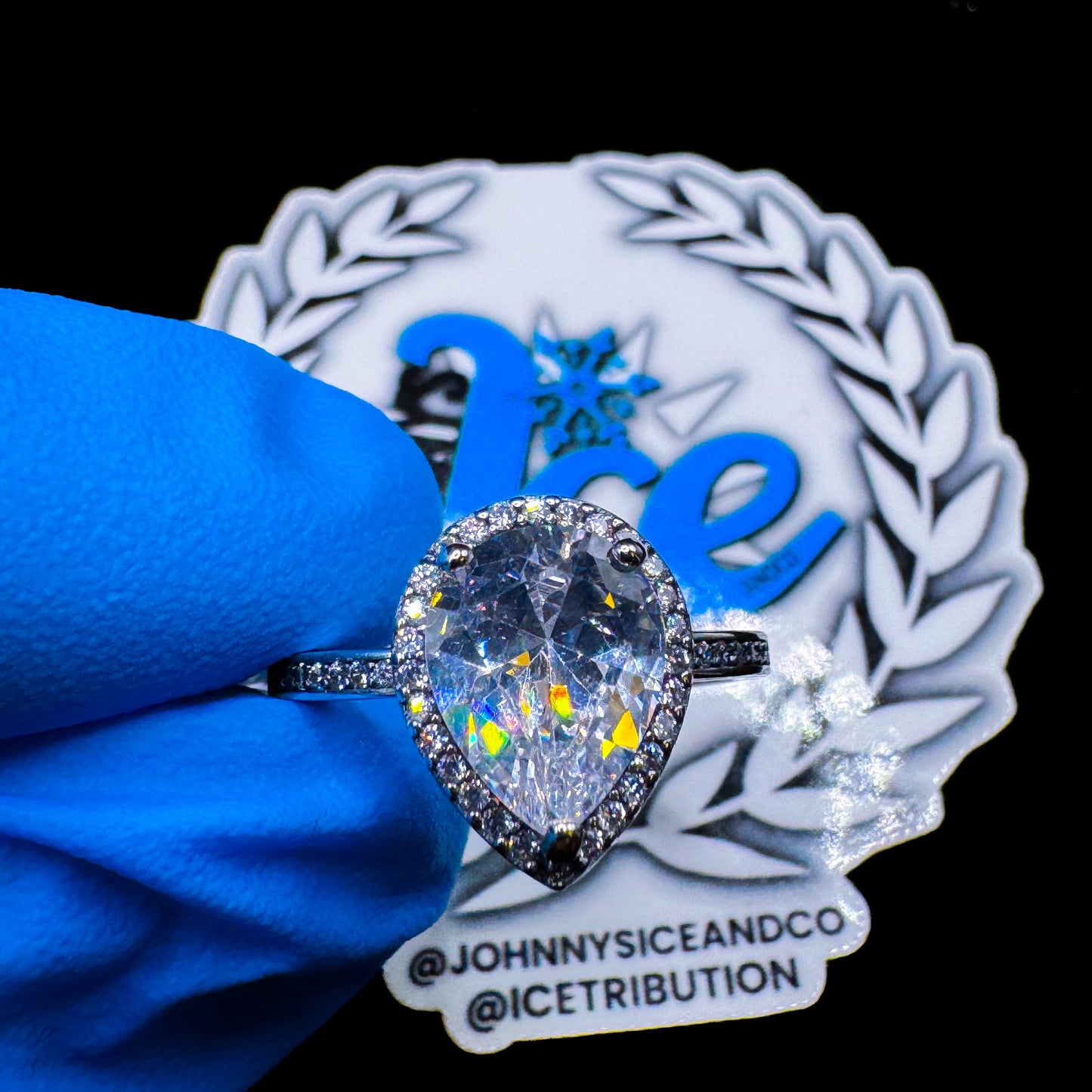 A teardrop-shaped silver band ring with a clear central stone and smaller stones around it, displayed on a blue cloth with a branded logo in the background.