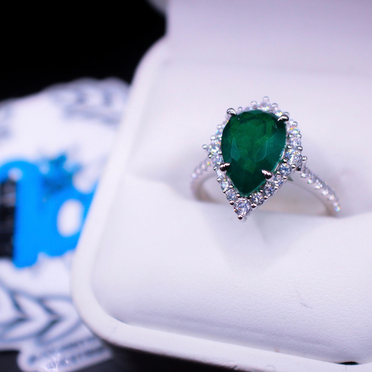 A teardrop-shaped emerald ring with a halo of smaller diamonds around it, set in a silver band, displayed in a white box with a blue and white pattern.