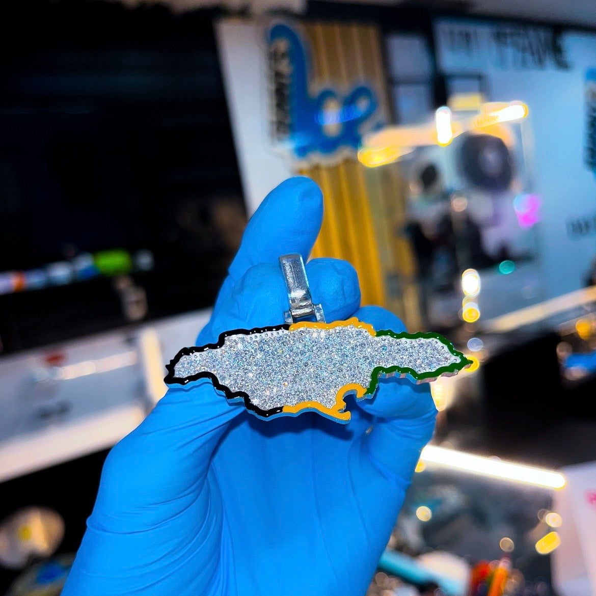 Hand wearing a blue glove holding a pendant shaped like a state, with a blurred indoor background.