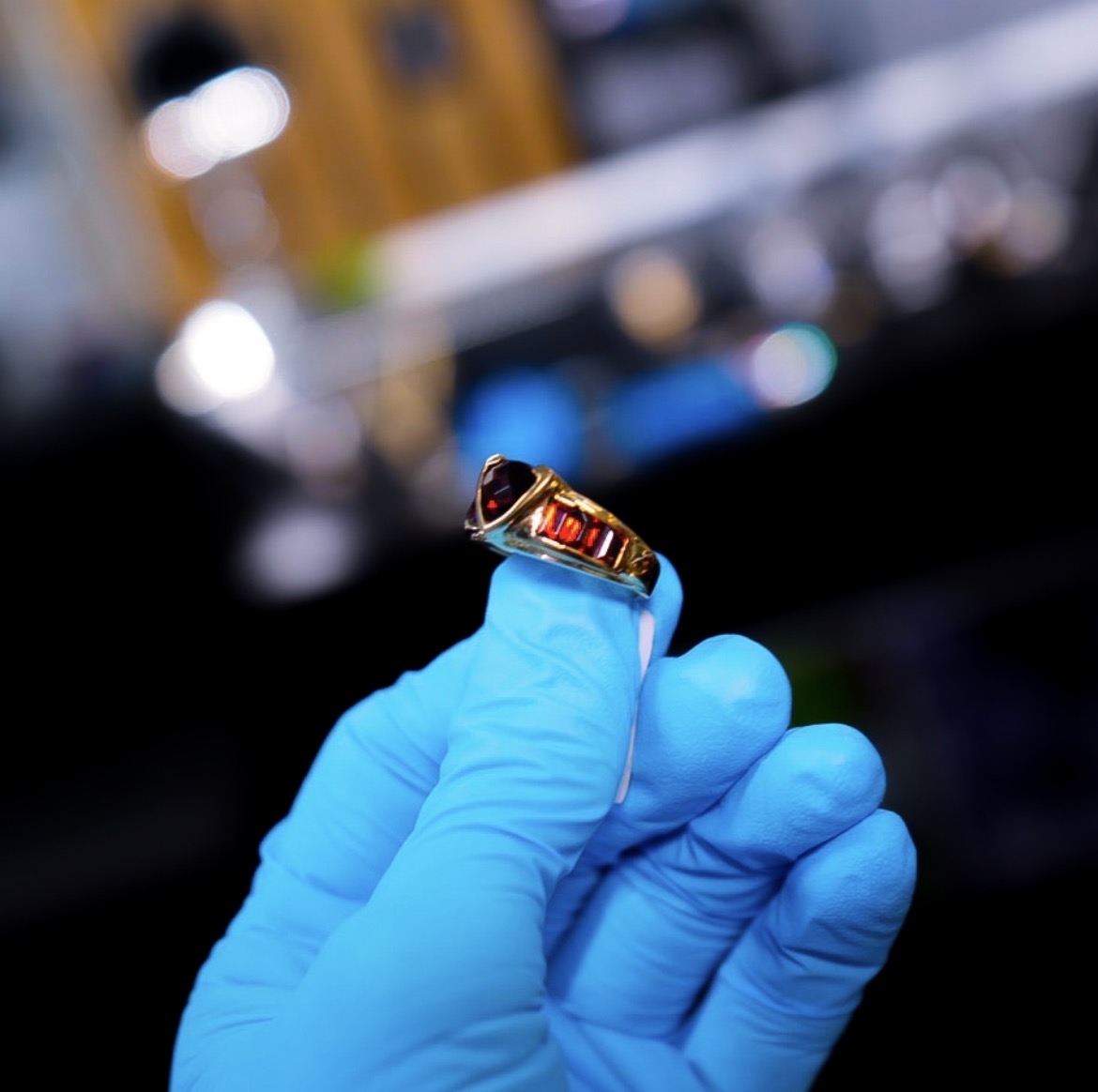 Gold ring with a red gemstone held by a gloved hand against a blurred background