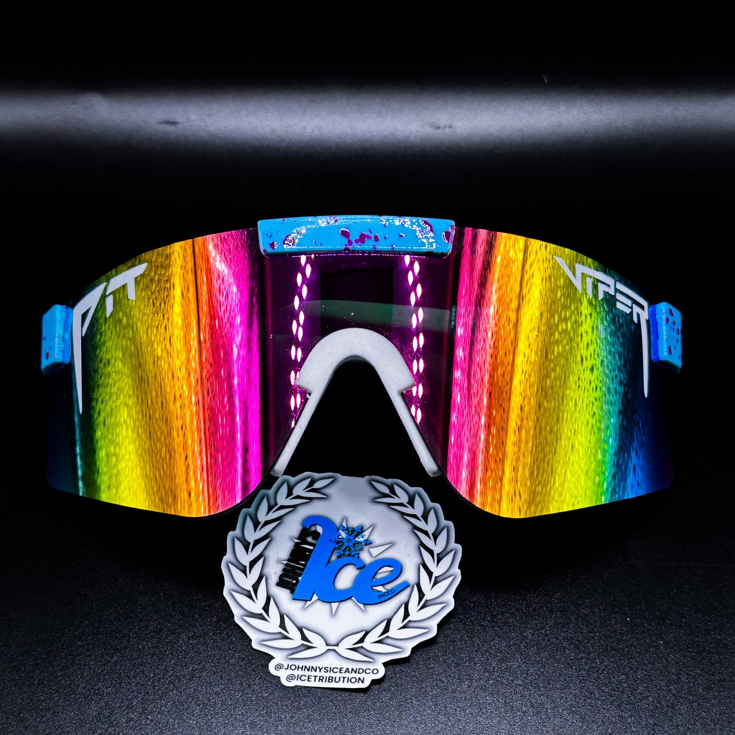 A pair of sunglasses with multicolored reflective lenses and pink/yellow frame, displayed with a logo and text on a reflective surface.