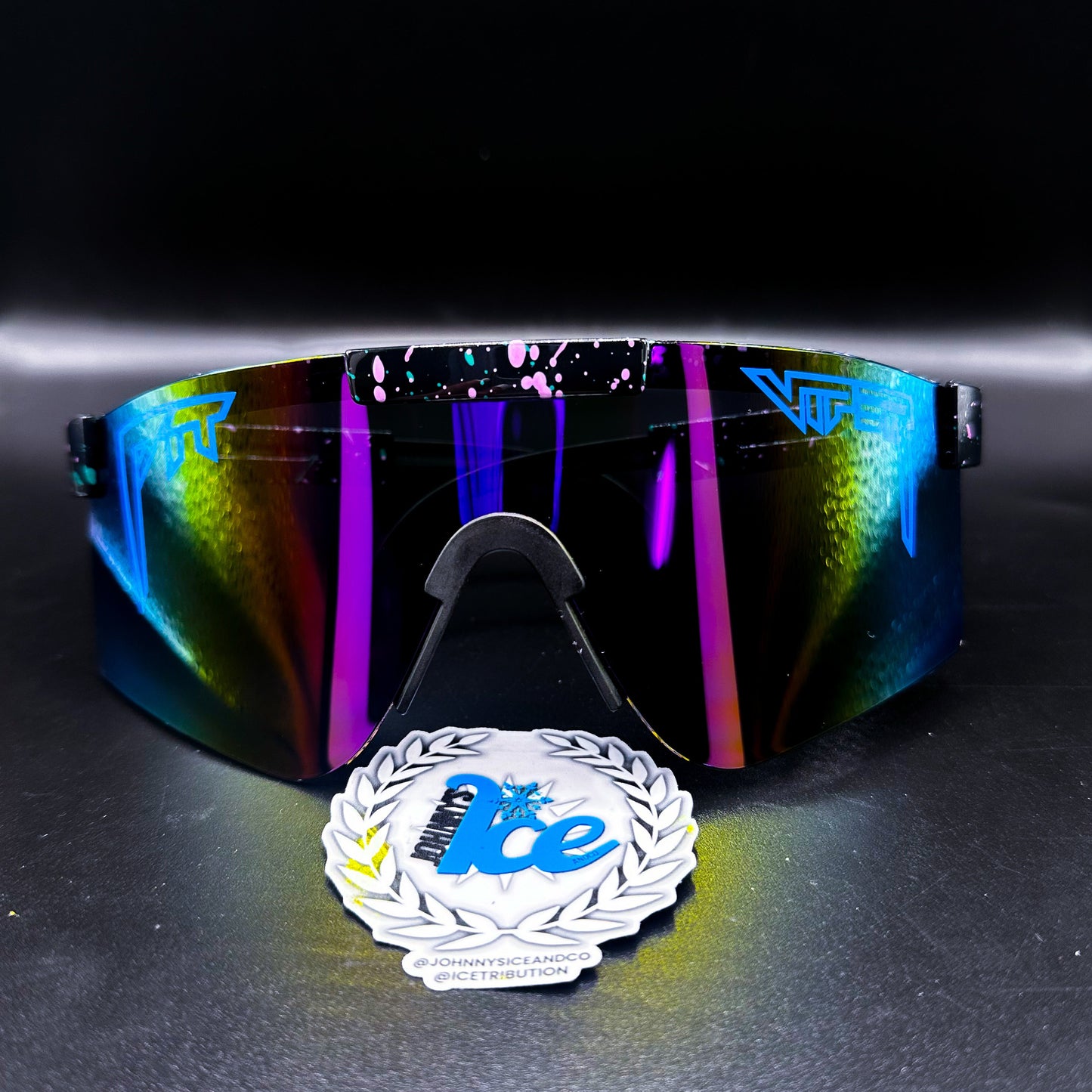 A pair of sunglasses with black and purple lenses, featuring a reflective multicolor design, and an award badge in front.