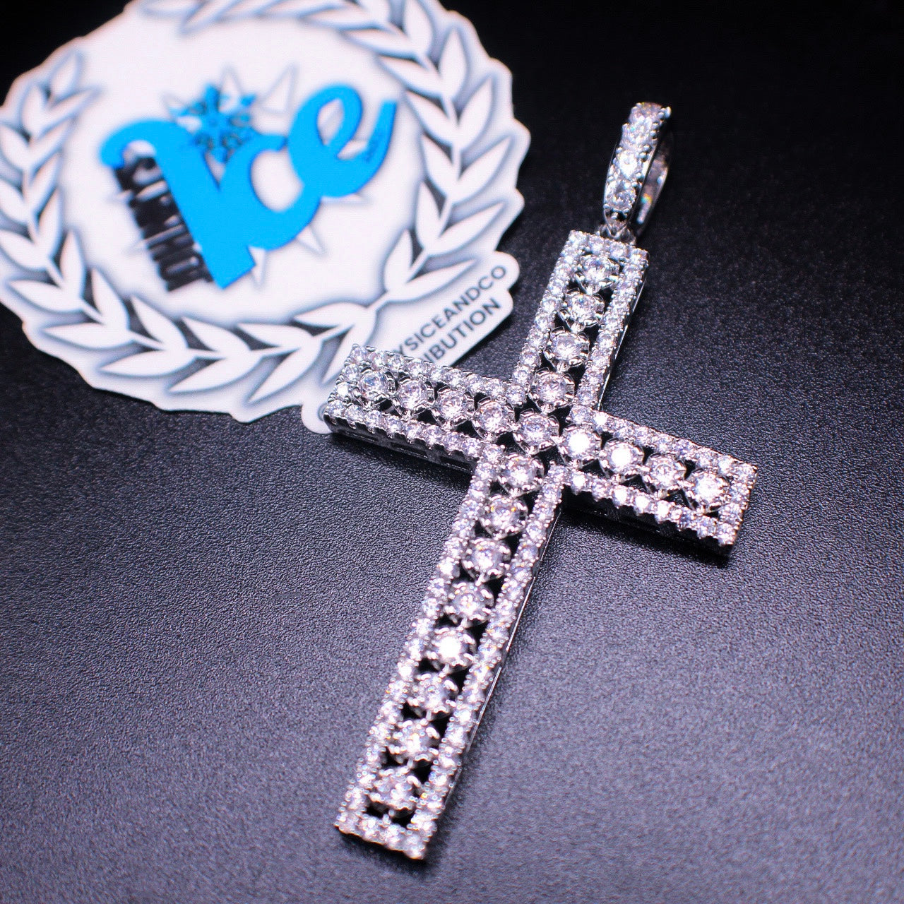 A silver cross pendant with a solitaire stone design, displayed against a dark background with a blurred logo in the top left corner.