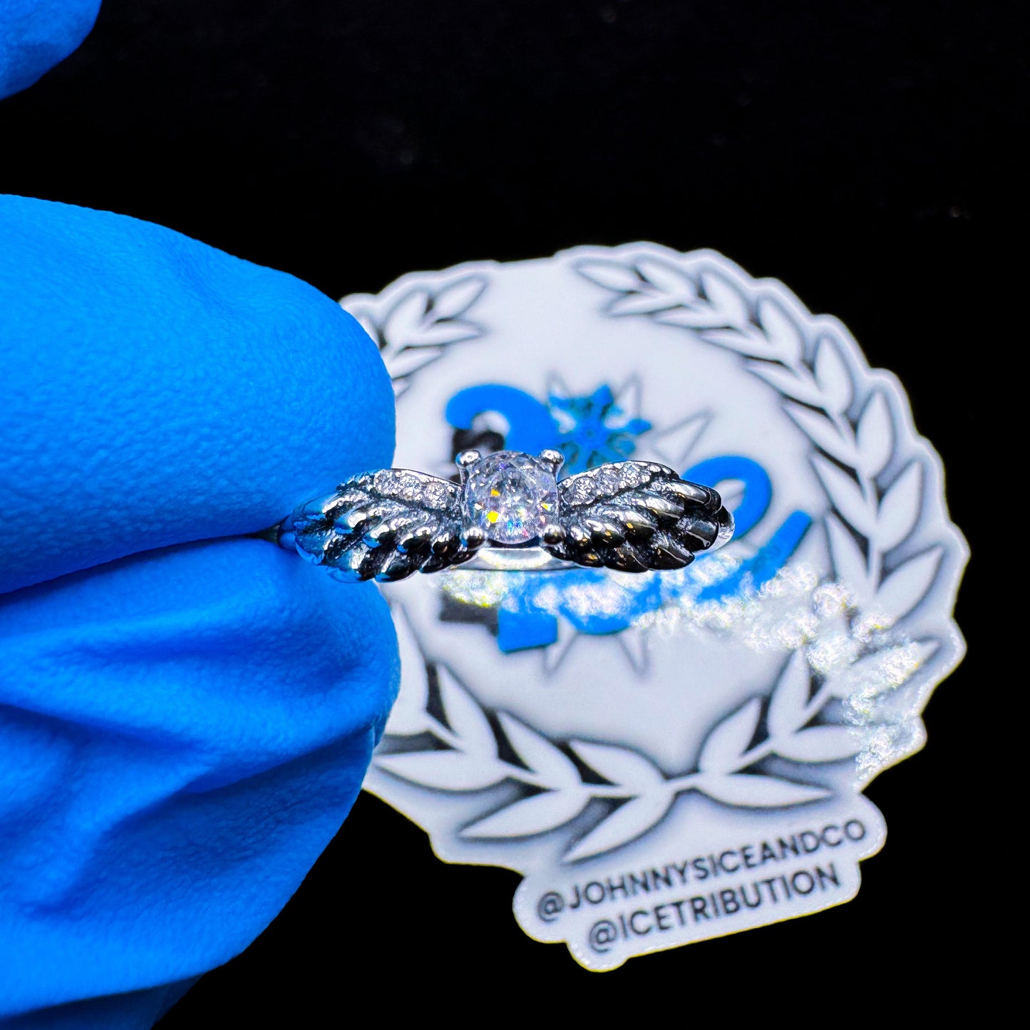 A silver ring with a wing design and a central cubic zirconia, displayed on a blue cushion with a branded backdrop.