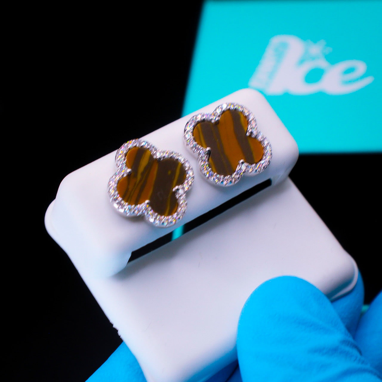 Clover-shaped earrings with brown center and silver border on a white stand, with a blue glove and box in the background.