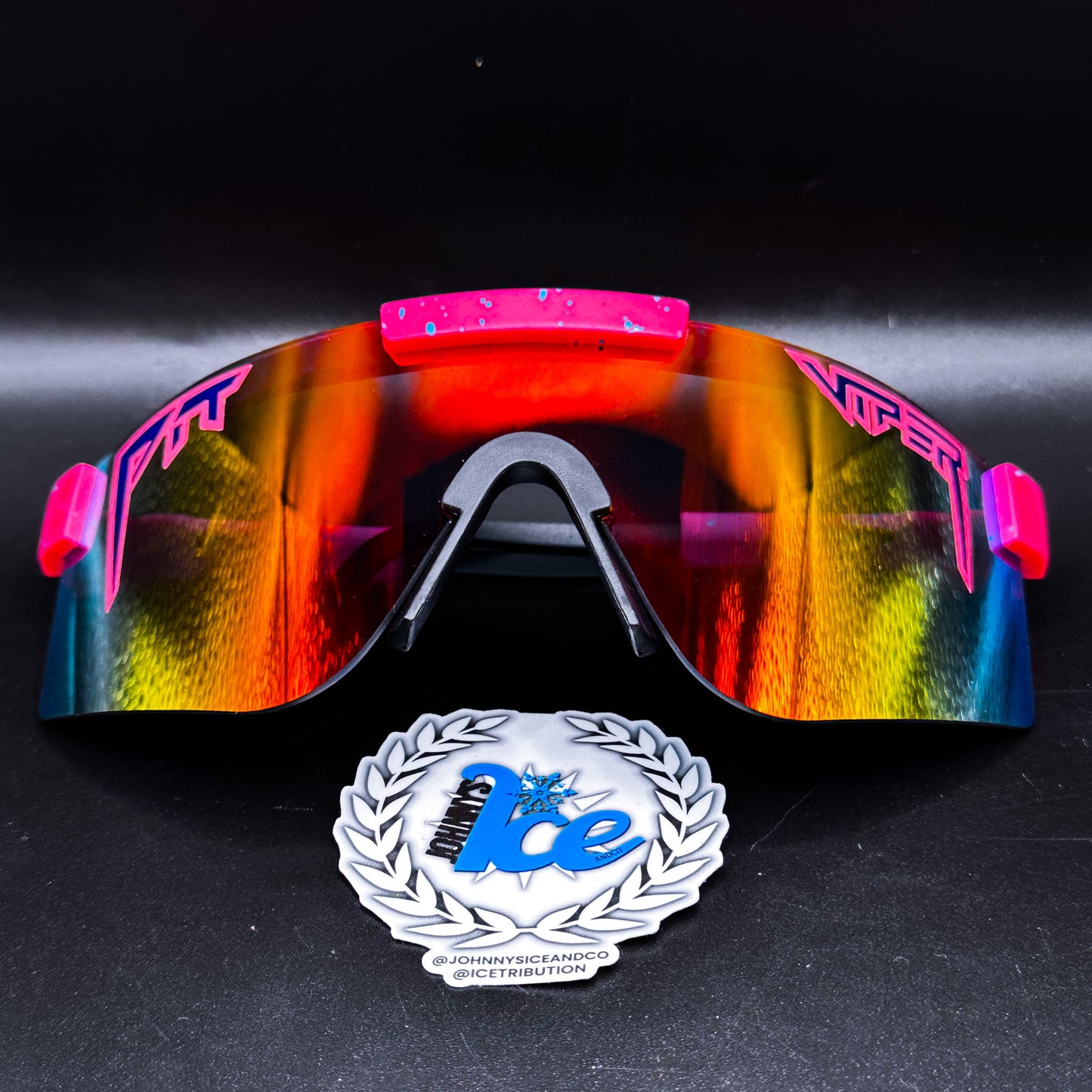 A pair of sunglasses with pink and yellow lenses, featuring a reflective surface and black frame, displayed with a logo sticker in front.