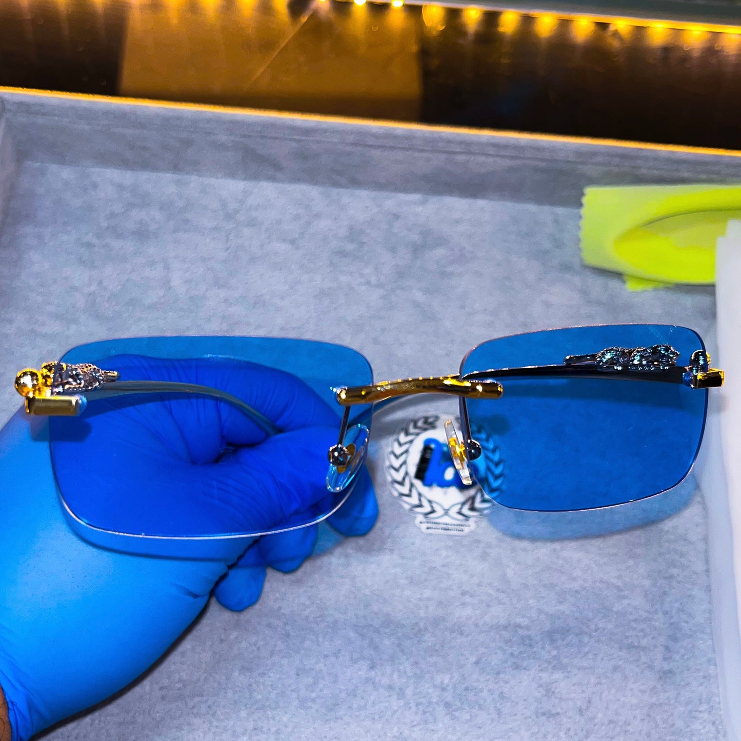 A pair of sunglasses with a blue tinted lens held by a gloved hand, with a light blue frame and decorative elements on the arms.