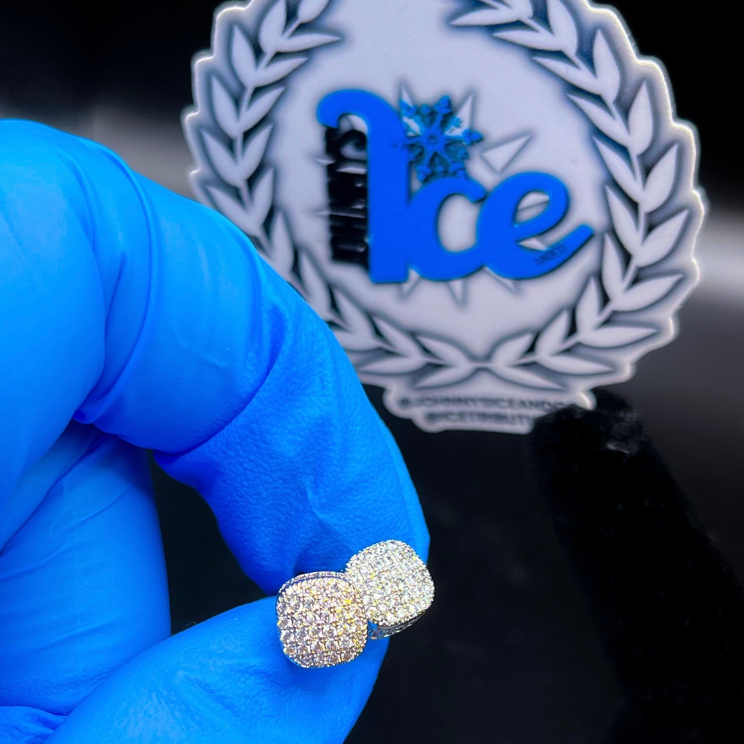 A pair of silver bubble-shaped earrings with cubic zirconia, held by a gloved hand, with a blurred logo in the background.