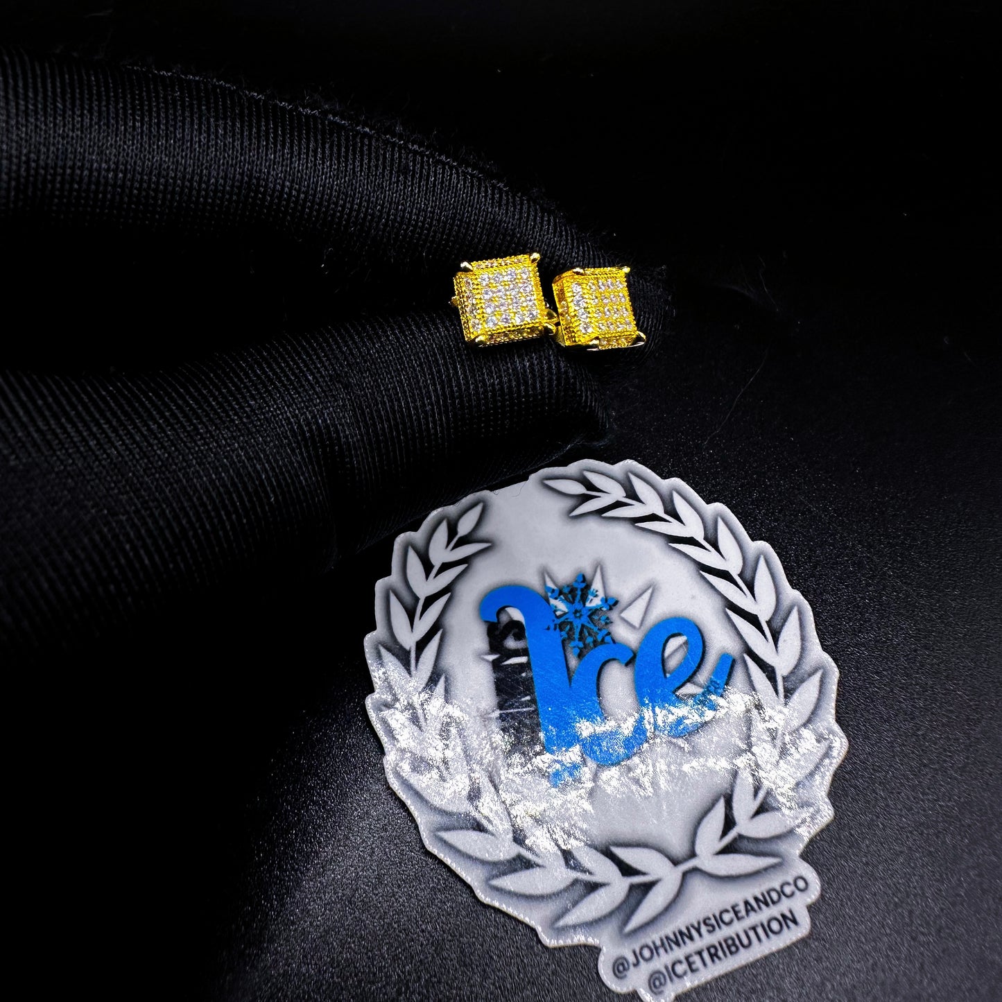 A pair of yellow gold screw-back earrings featuring cubic zirconia, displayed on a black surface next to a decorative emblem.