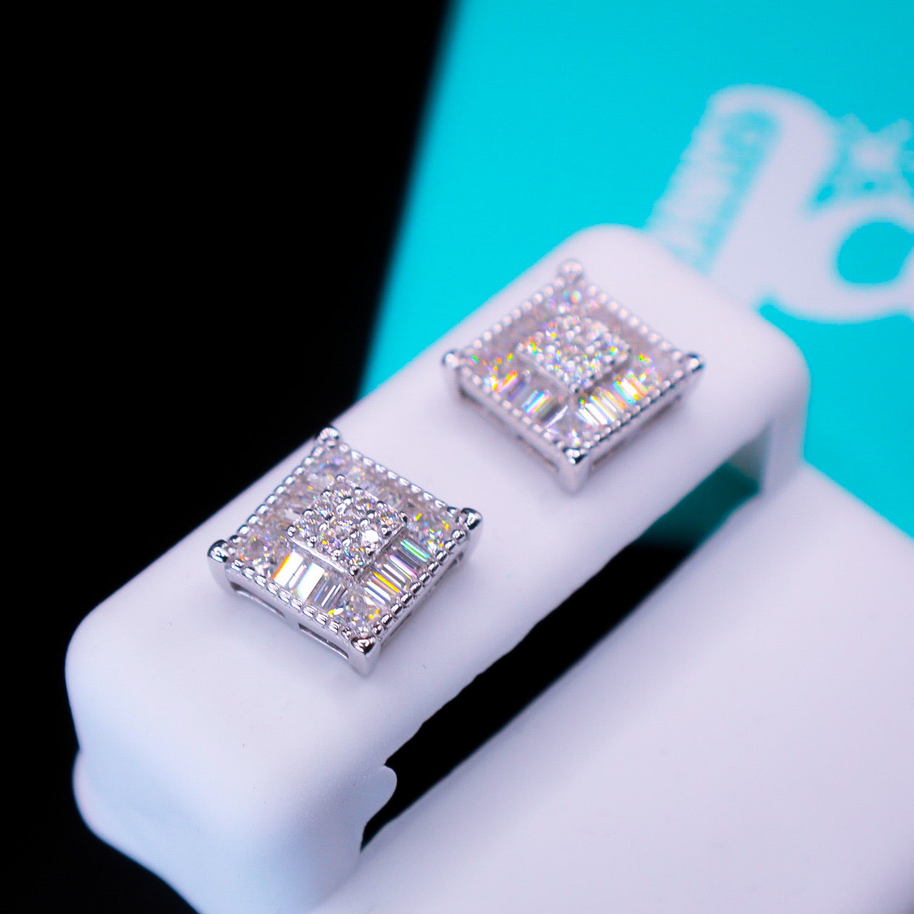 Square-shaped silver earrings with clear gemstones on a white stand against a black background.