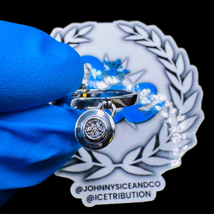 A silver ring with a clear central gemstone, possibly a diamond, surrounded by smaller clear stones, displayed against a branded background.