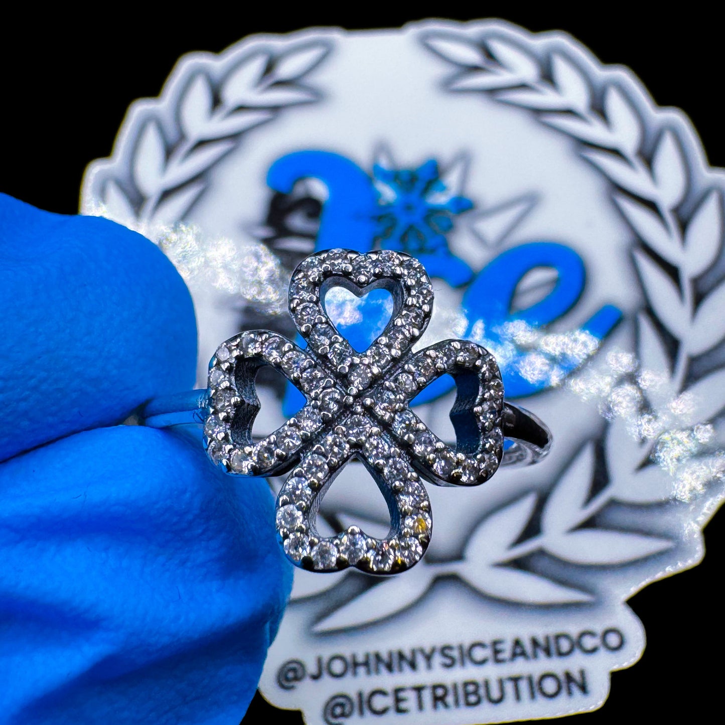 A silver ring with a clover shape and heart cutouts, adorned with clear cubic zirconia stones, displayed on a blue cushion.