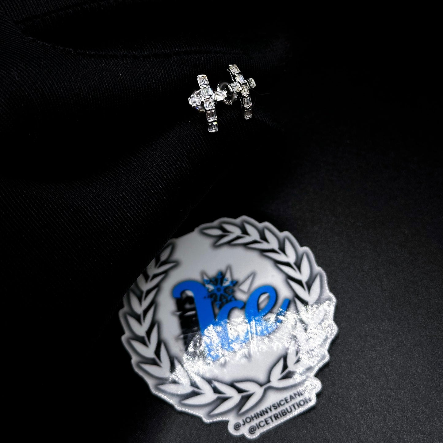 A pair of silver cross earrings with cubic zirconia displayed on a black surface, accompanied by a branded round box.