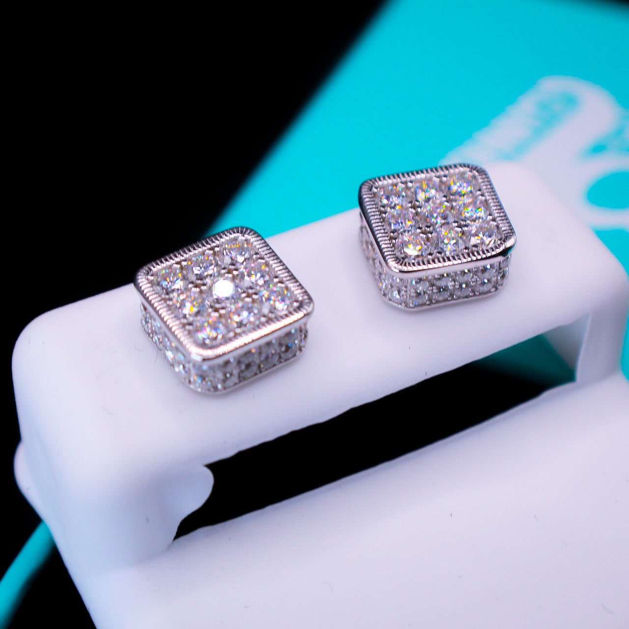 Square-shaped silver earrings with clear gemstones on a white stand.