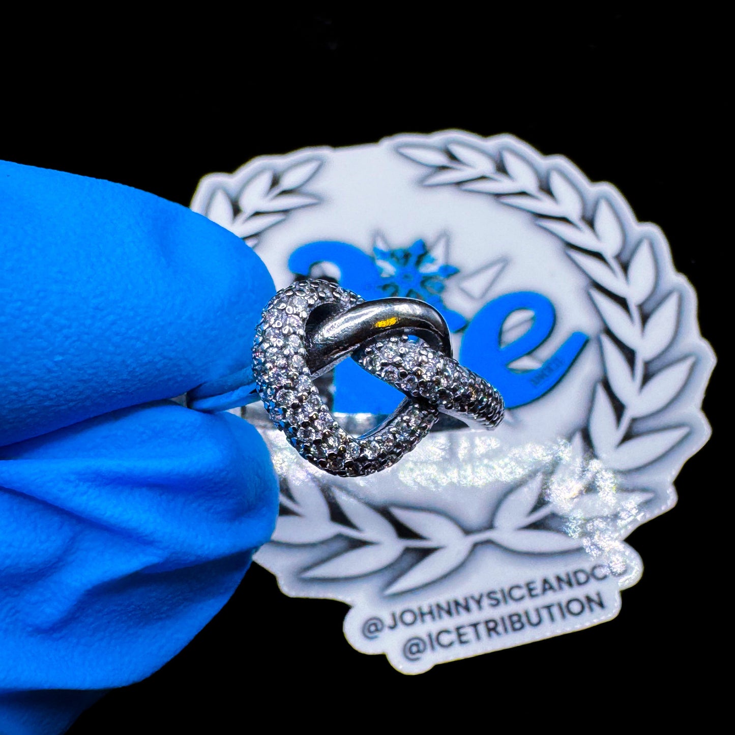 A entangled heart-shaped ring with clear cubic zirconia stones, displayed in front of a brand-related emblem with a blue gloved hand holding it.