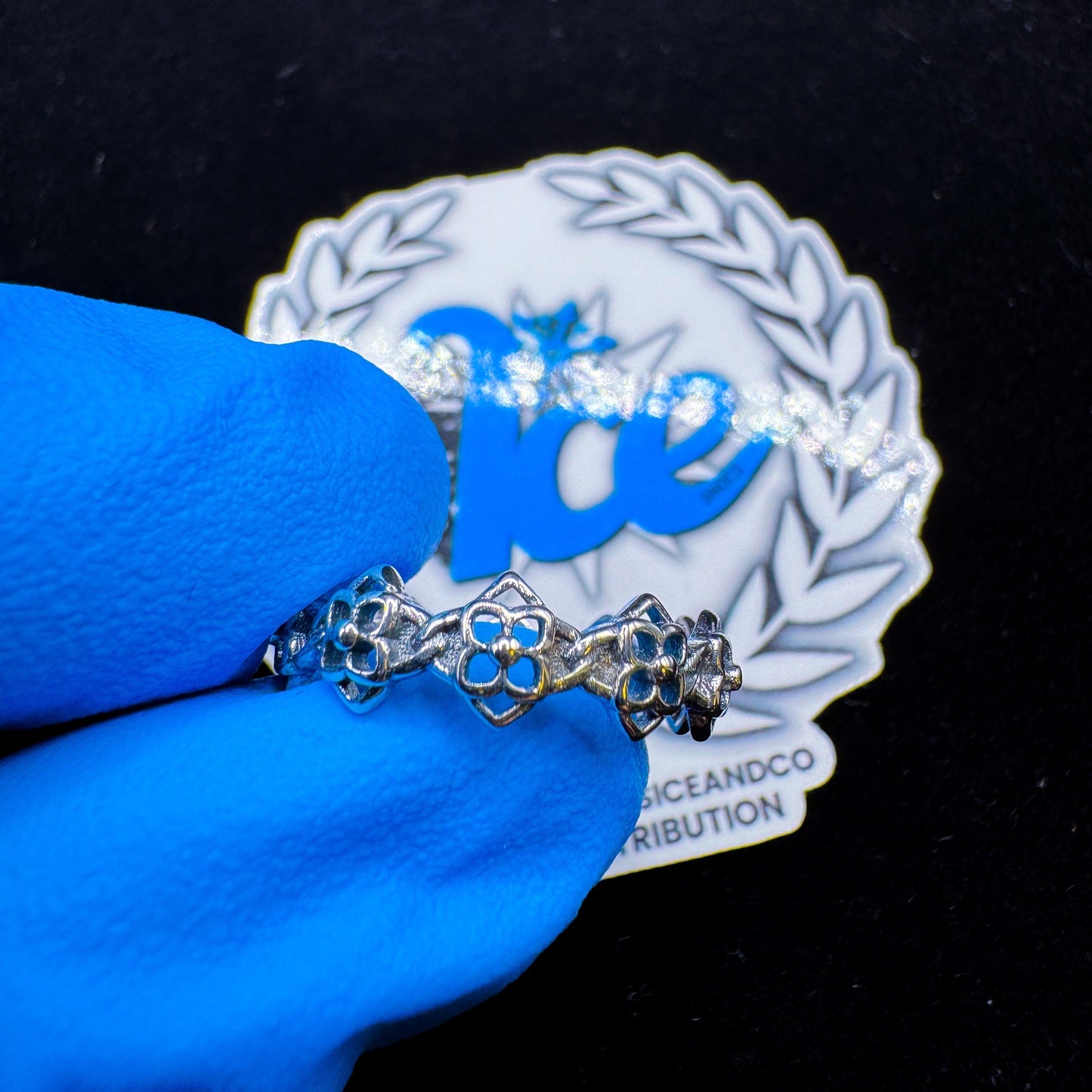 A silver ring with a floral design, displayed on a blue gloved hand with a brand logo in the background.
