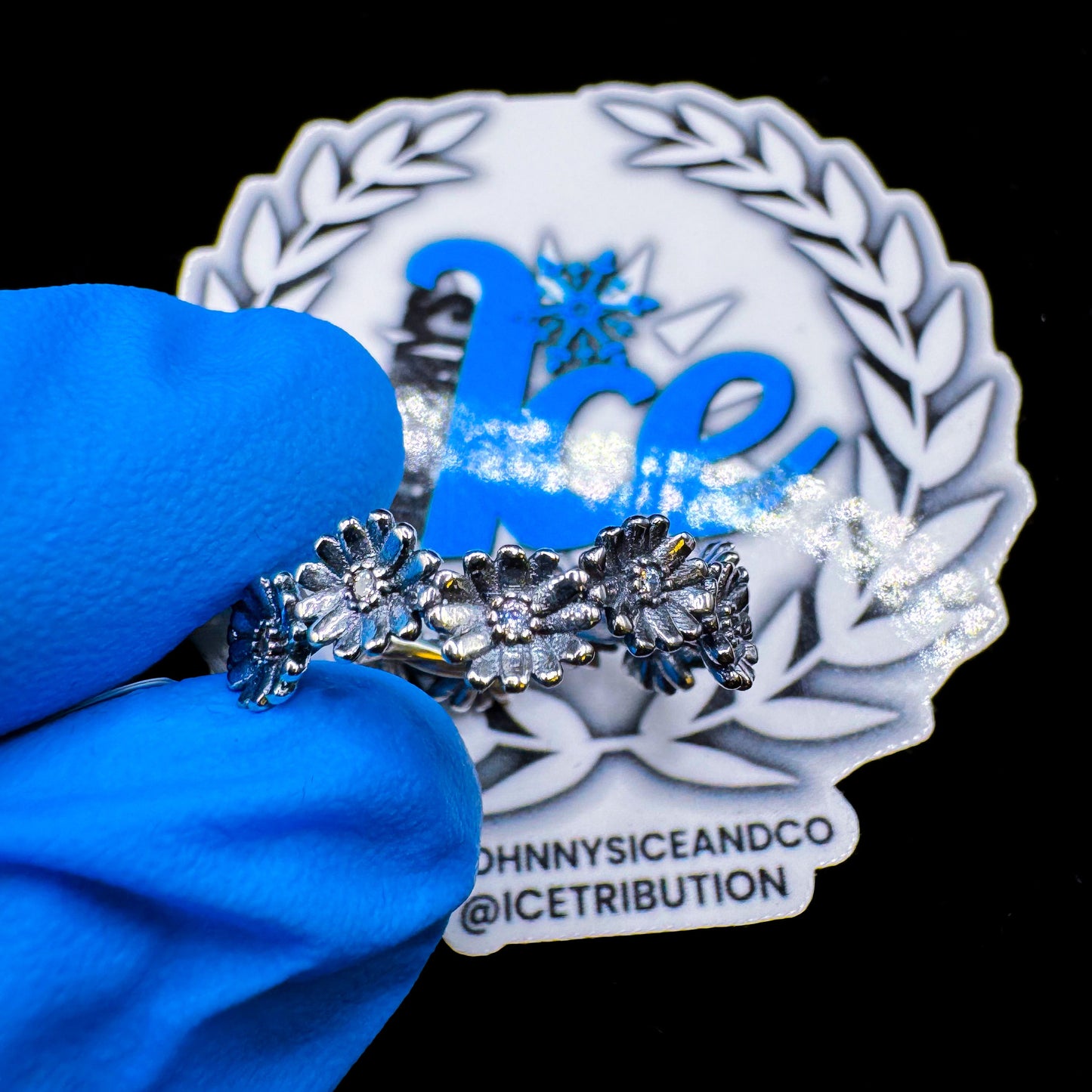 A silver ring with a floral design, featuring multiple clear cubic zirconia stones, held by a blue gloved hand, with a branded backing plate behind.
