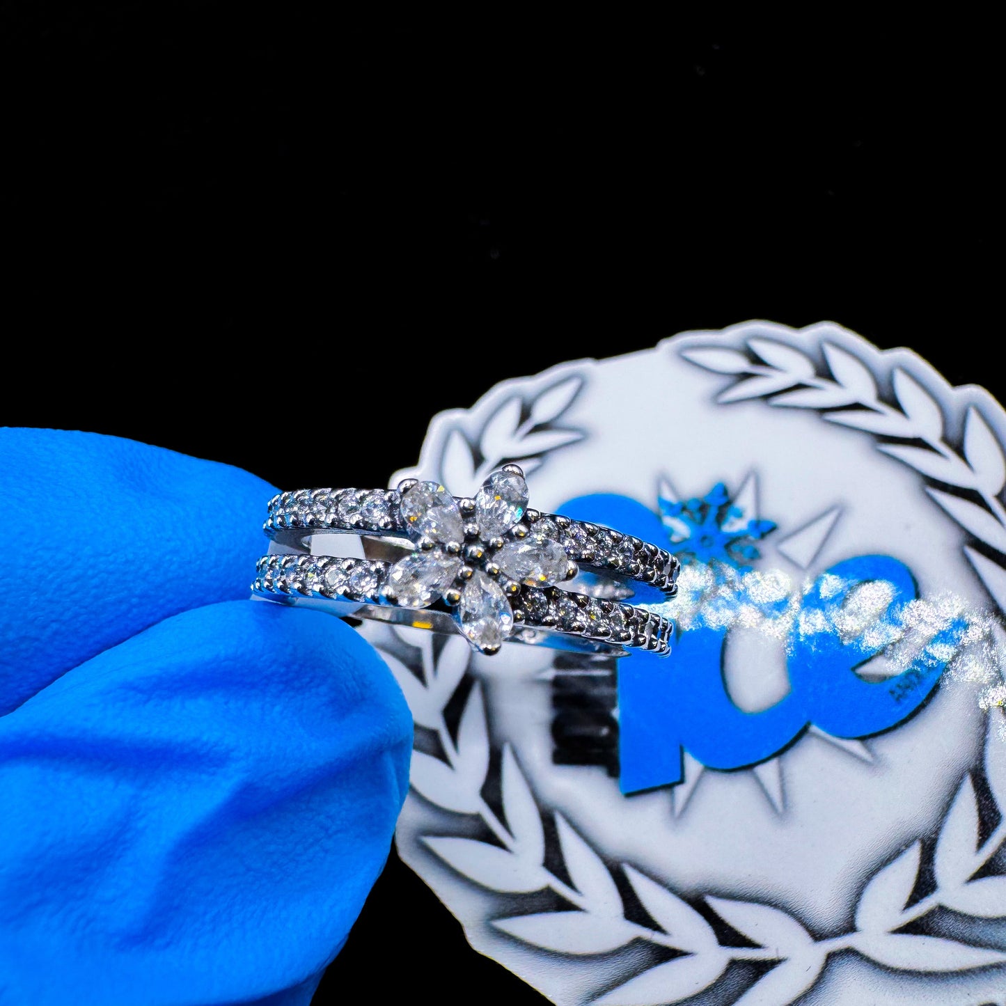 A silver ring with two bands of flowers and multiple clear gemstones, displayed on a blue cushion.