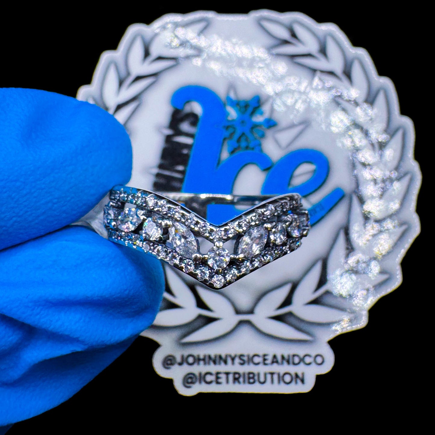 A silver garter ring with multiple clear cubic zirconia stones set in a curved design, displayed on a blue gloved hand with a branded logo in the background.