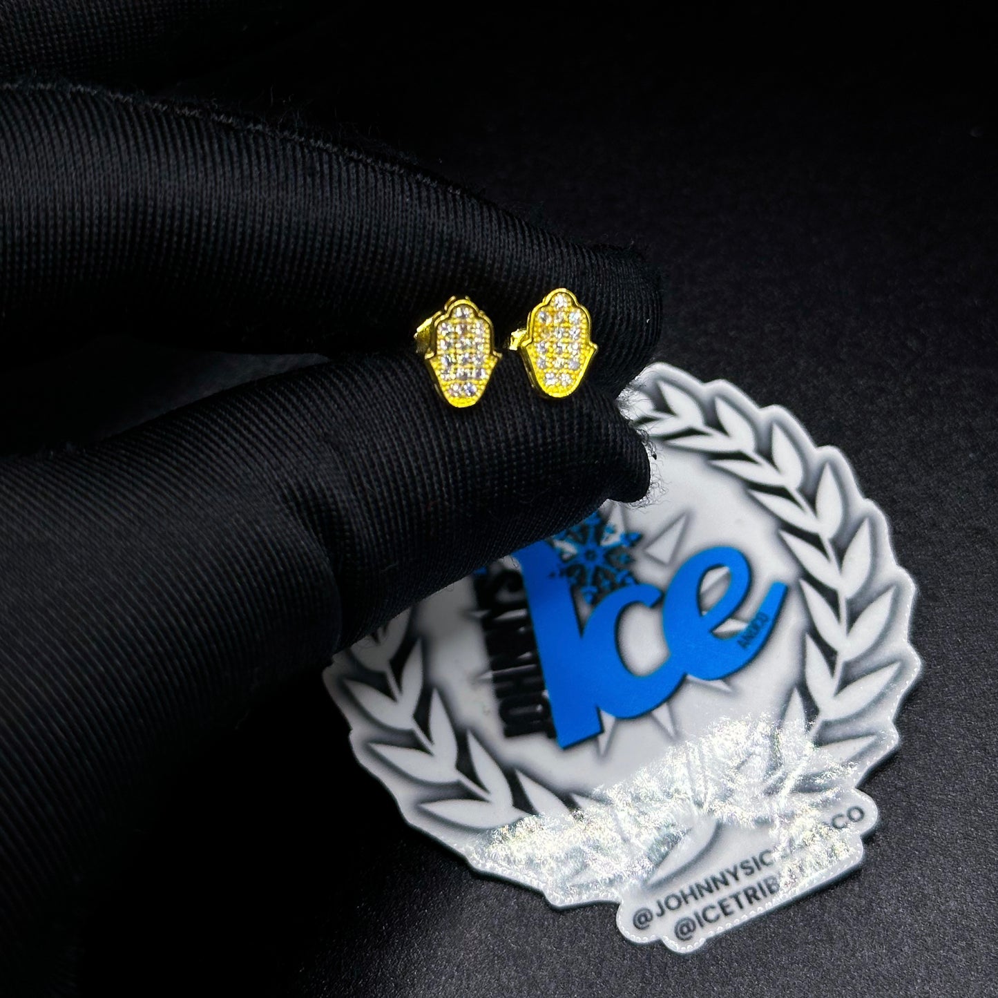 A pair of yellow gold Hamsa earrings with diamond-like studs on a black surface, with a reflective metallic sign in the background.