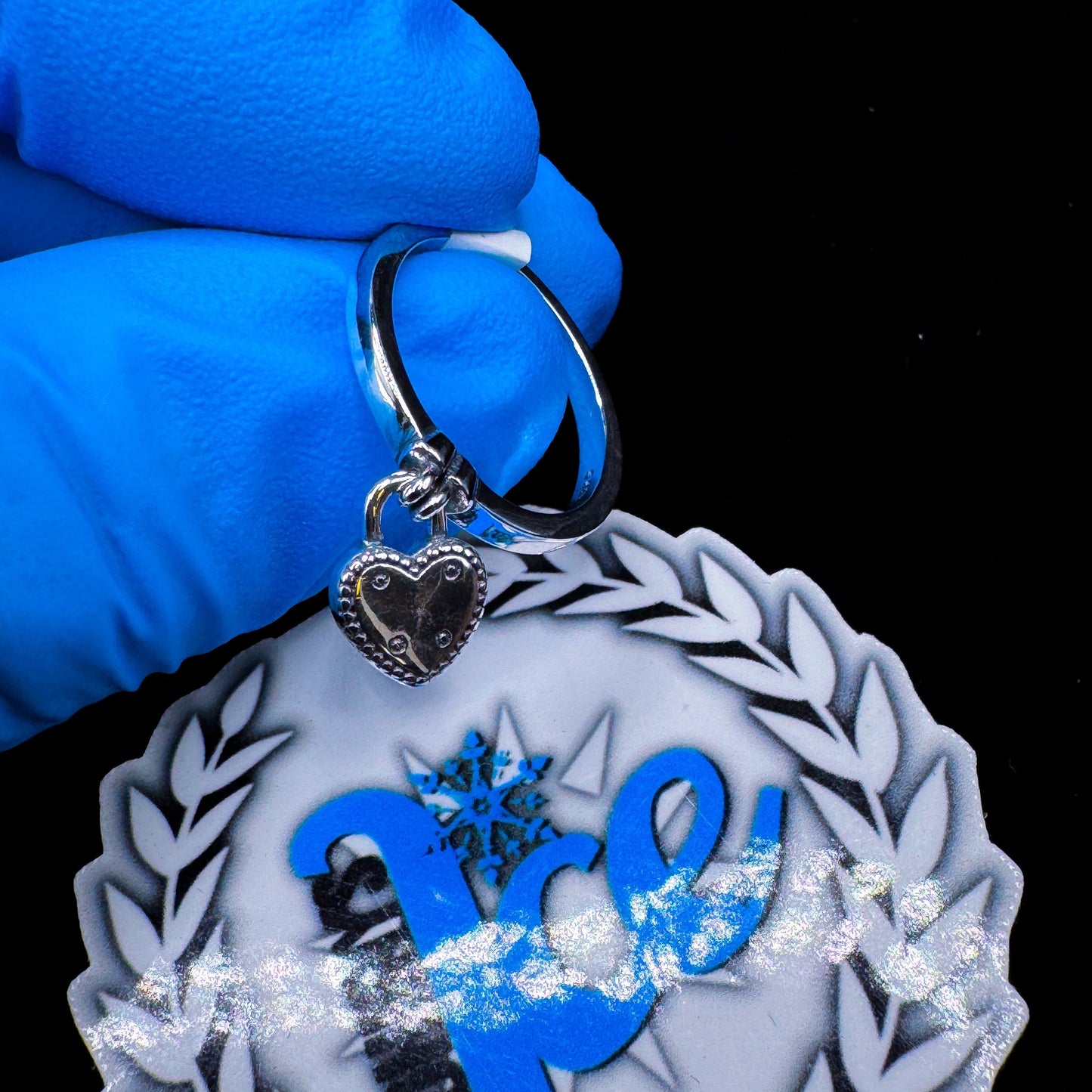 A silver heart-shaped locket attached to a ring, displayed against a black background with a blue-gloved hand holding the item.