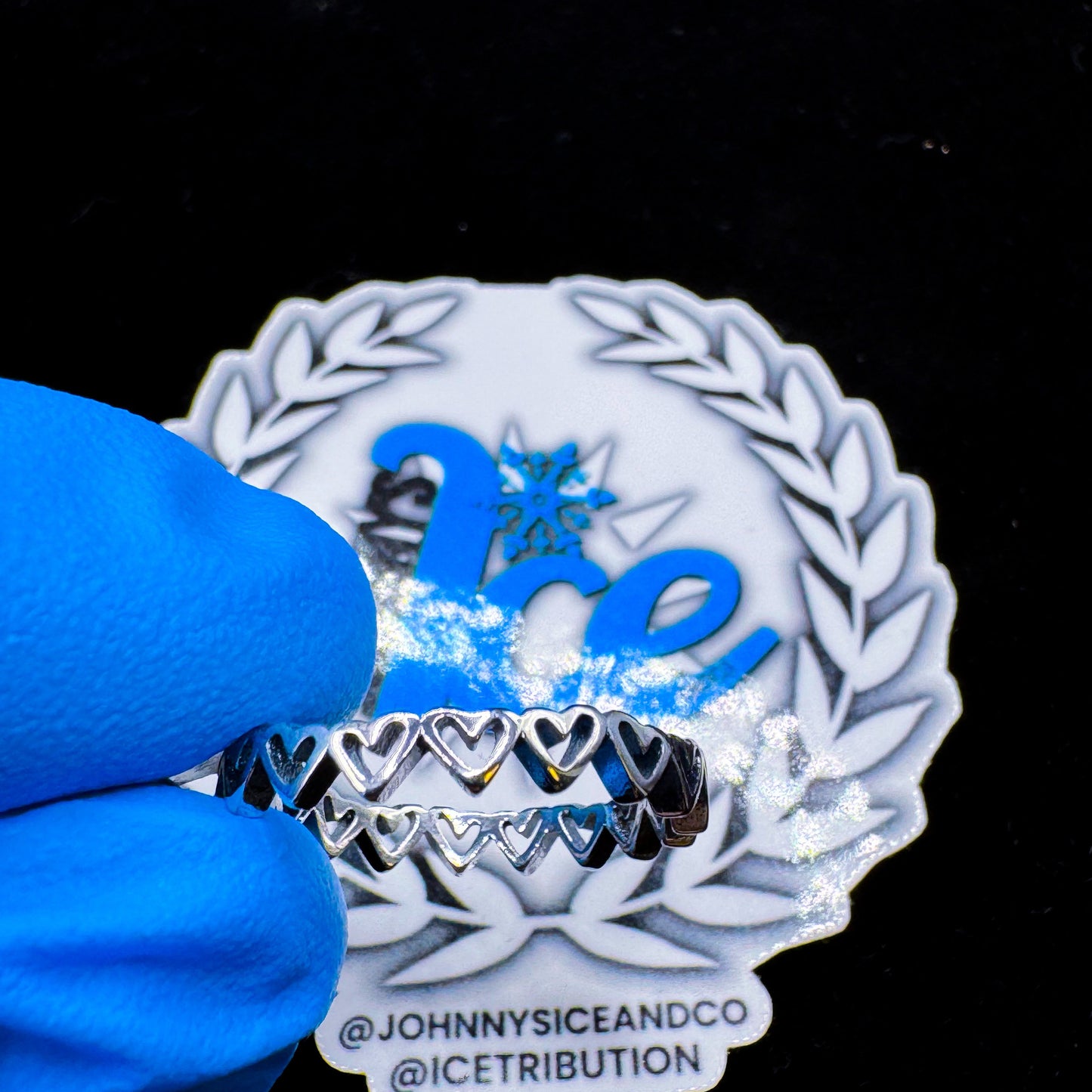 A silver ring with heart-shaped cutouts and intricate designs, held by a blue gloved hand, with a branded backing plate.