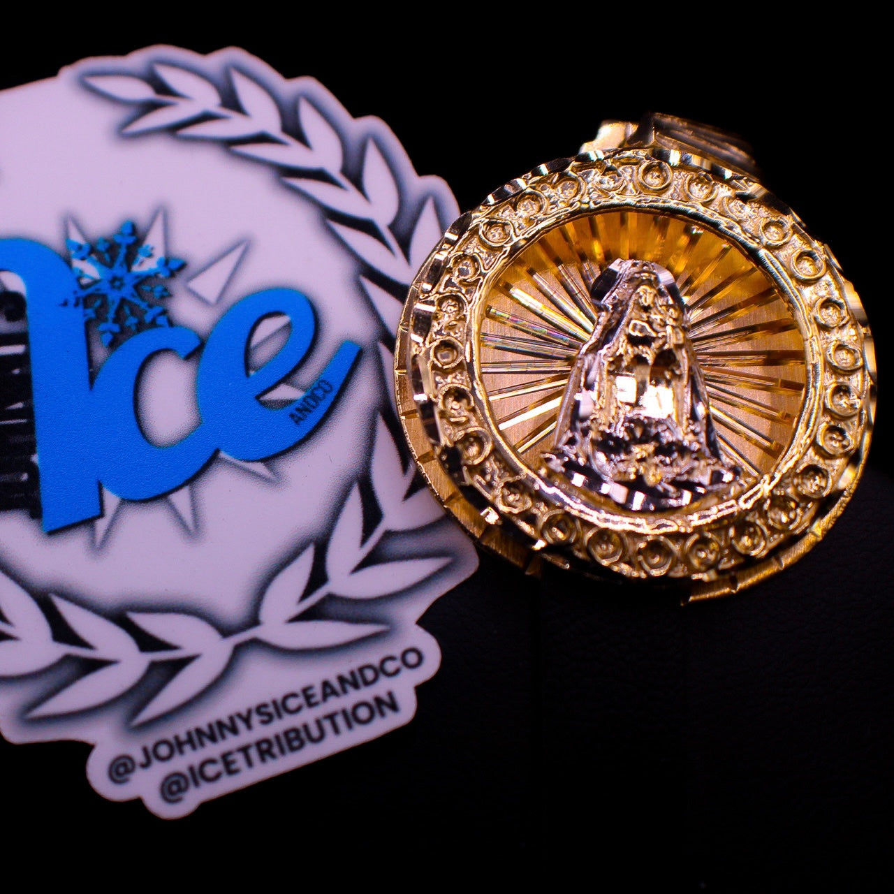 A gold medallion featuring an image of Mother Mary with intricate detailing, displayed against a black background with a reflection below.