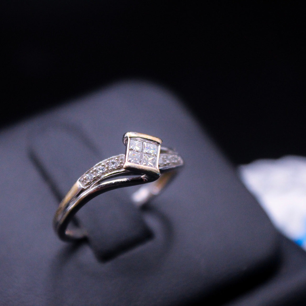 A princess cut diamond ring with a 10K white gold band, displayed on a black surface, possibly in a jewelry case.