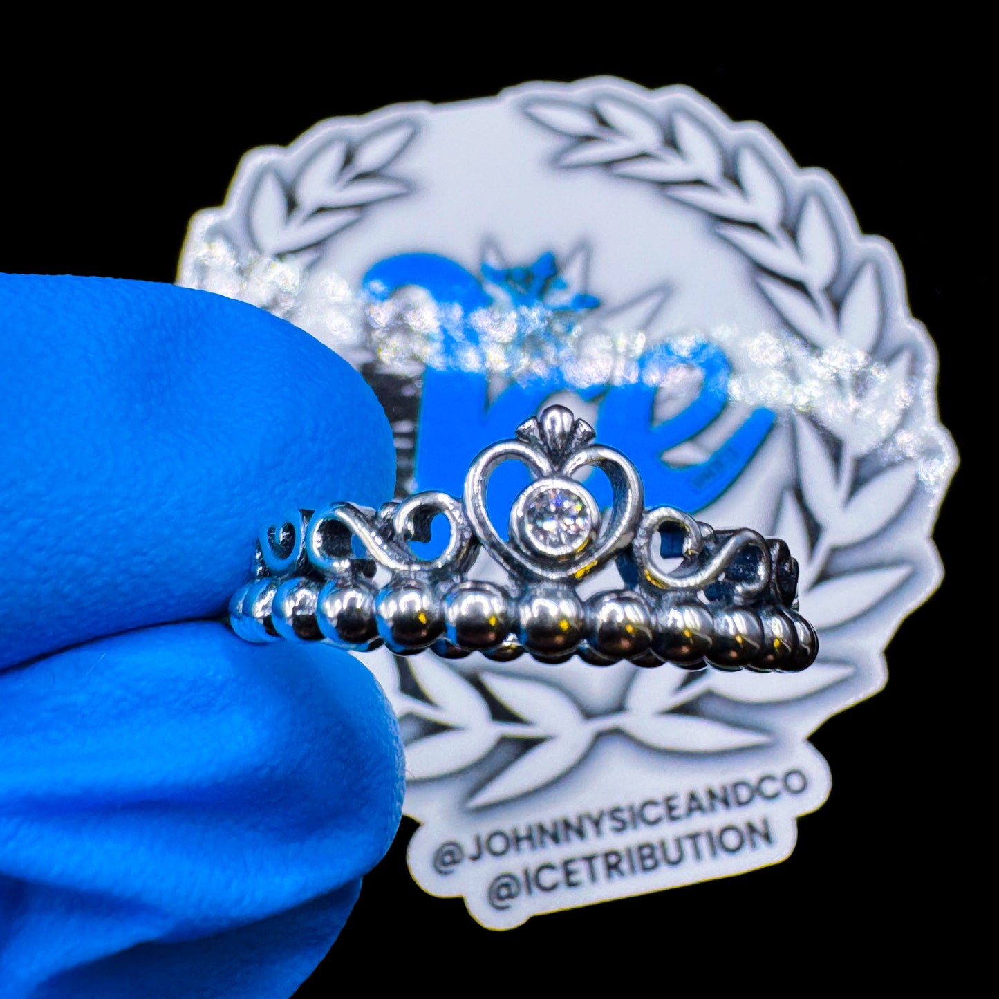A sterling silver princess heart crown ring with cubic zirconia, displayed on a blue cloth with the brand logo visible in the background.