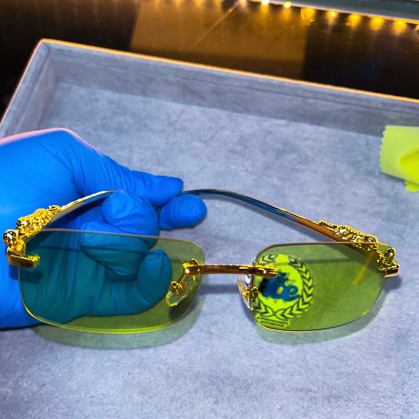 A pair of sunglasses with light green lenses and gold-colored frames, displayed on a platform with a blue gloved hand beside it.