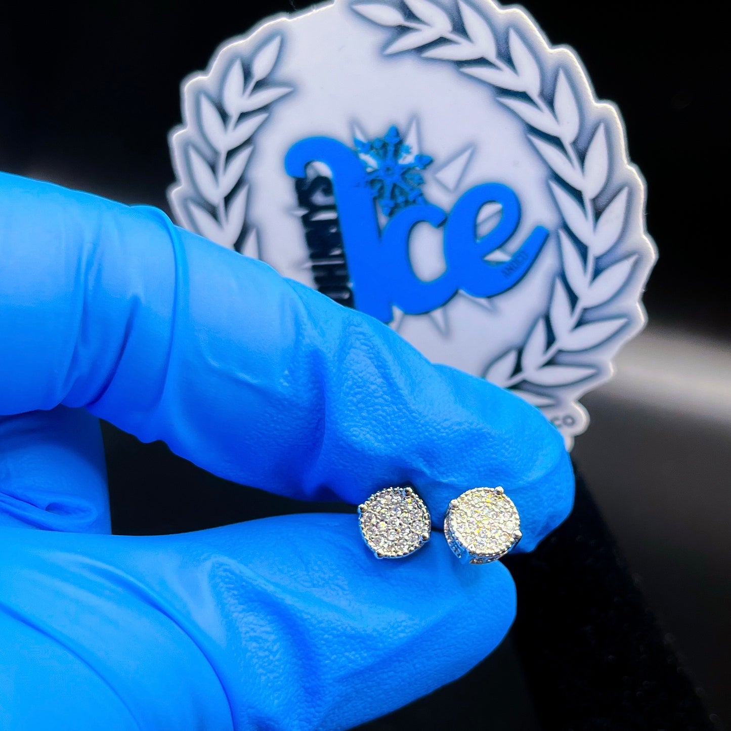 A pair of silver round barrel earrings with cubic zirconia, displayed in front of a brand-related emblem.