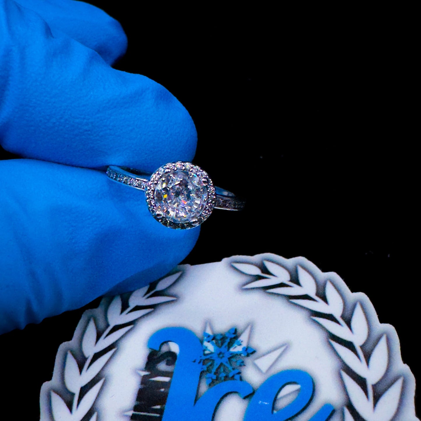 A silver ring with a round central diamond surrounded by smaller diamonds, held by a gloved hand, with a logo in the background.