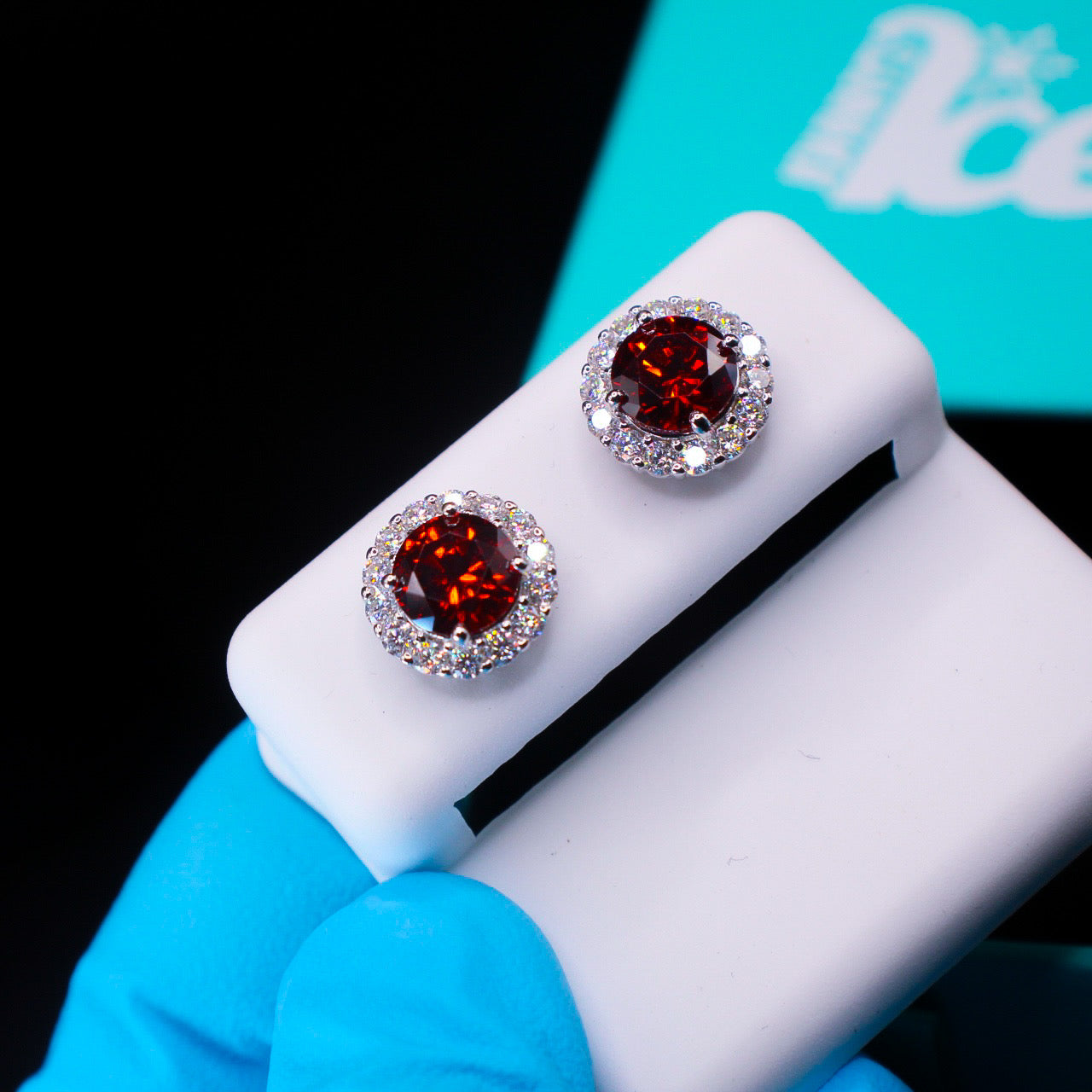Pair of red gemstone earrings on a white stand with a blue glove and turquoise box in the background.