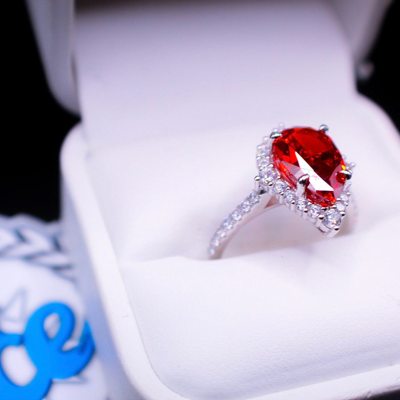 A teardrop-shaped red synthetic ruby ring with a halo of smaller clear stones, set in a silver band, displayed in a white box with a blue and white cloth around it.