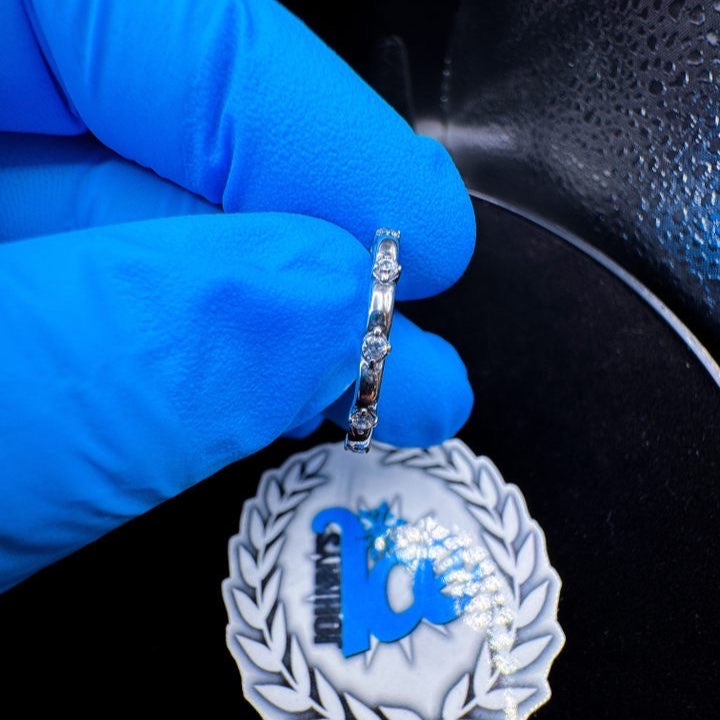 A person wearing a blue glove is holding a silver ring with multiple small diamonds or cubic zirconia stones.