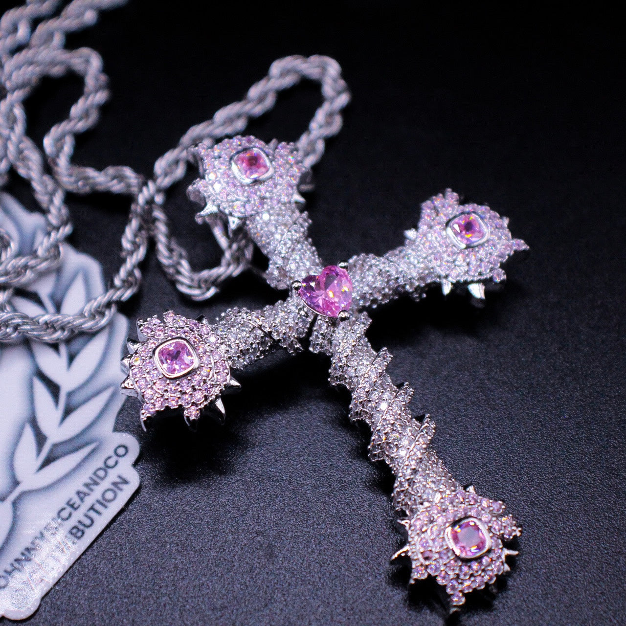 Silver finish cross pendant with pink heart shaped gem in center and clear diamonds all around laying on a black display