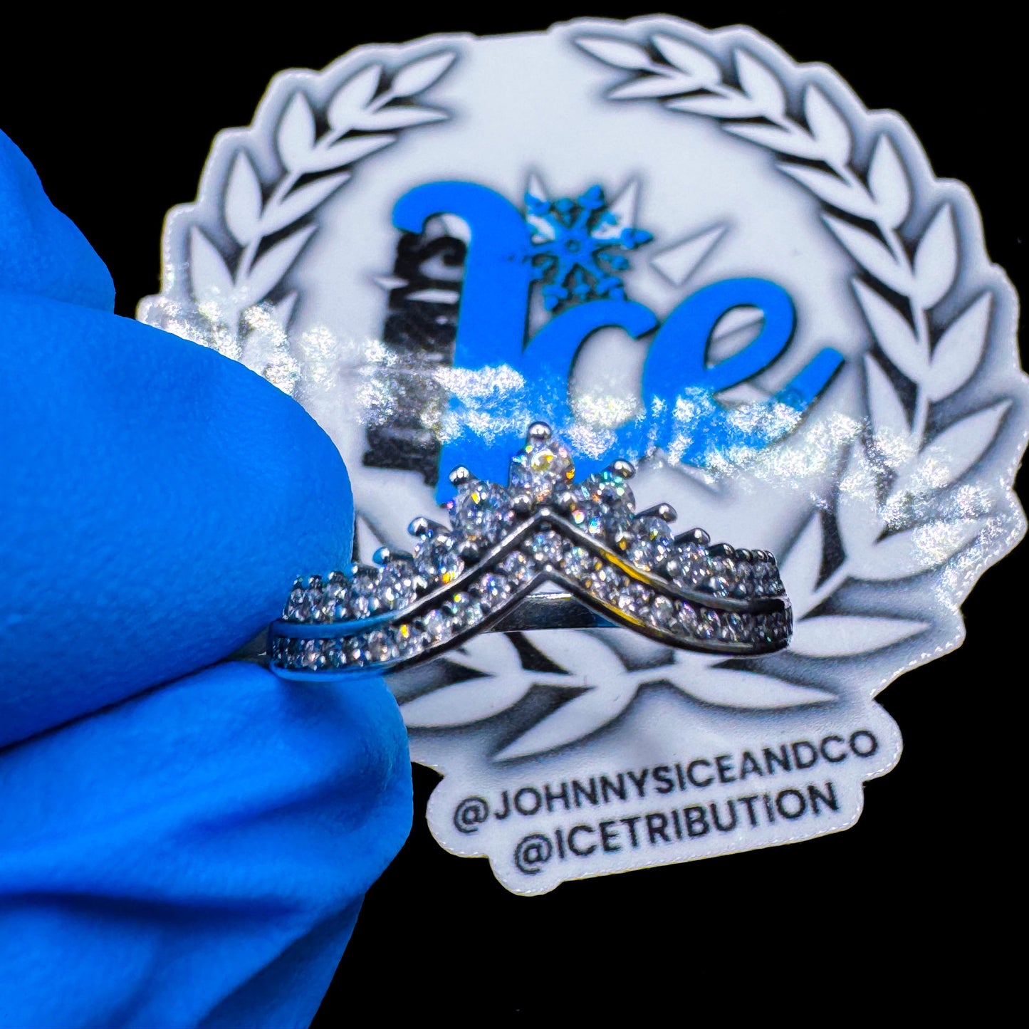 A silver-colored crown-shaped ring with blue and white accents, displayed on a blue cloth with the brand name 'Johnny's Ice & Co' visible in the background.