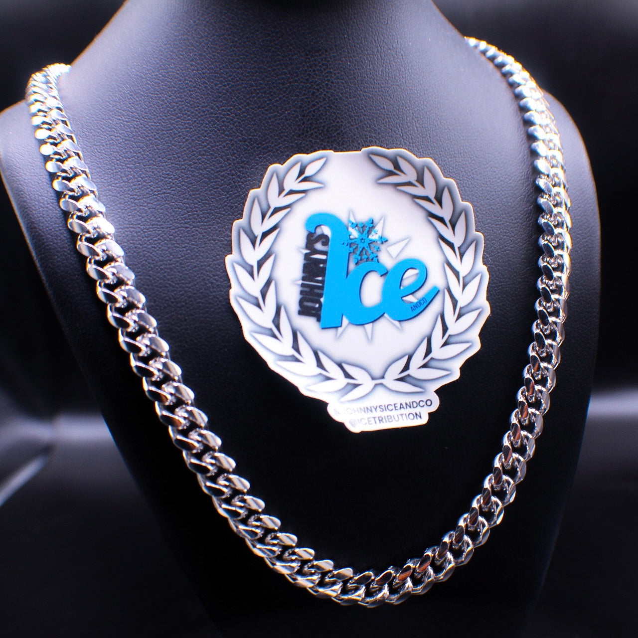 A silver Cuban link chain necklace displayed on a mannequin bust with a branded pin.
