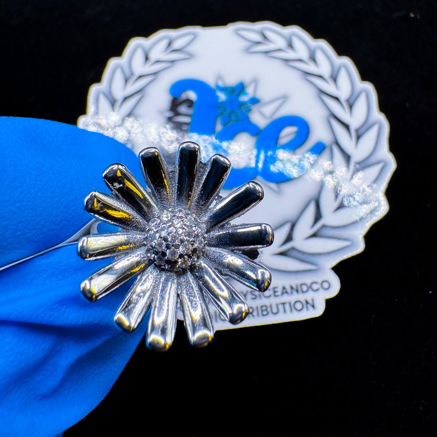A silver-colored floral ring with multiple petals, displayed on a blue fabric surface with a blurred logo in the background.