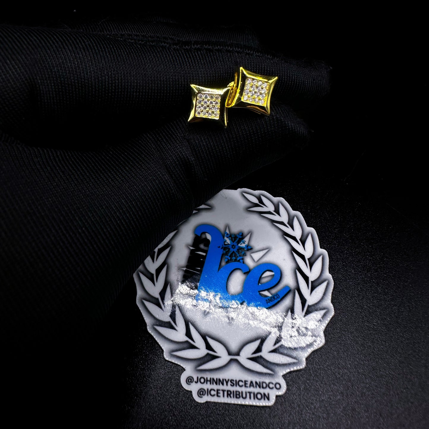 A pair of gold-colored square earrings with screw-back closures, displayed on a black background with a logo that includes the text 'Ice' and the name 'Johnny Ice & Co'.