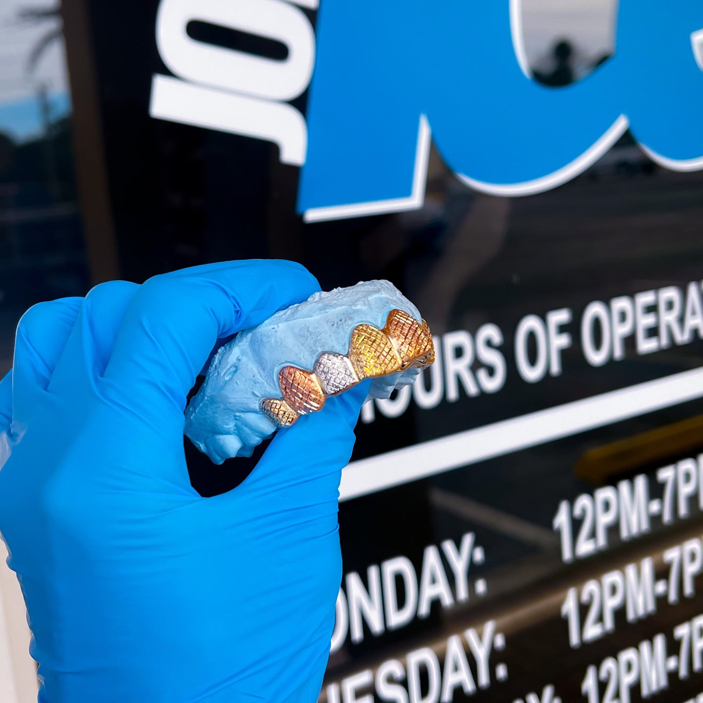 A set of tri-tone gold grillz held in a person's hand, with a blue glove on, in front of a sign that reads 'JIAC'.