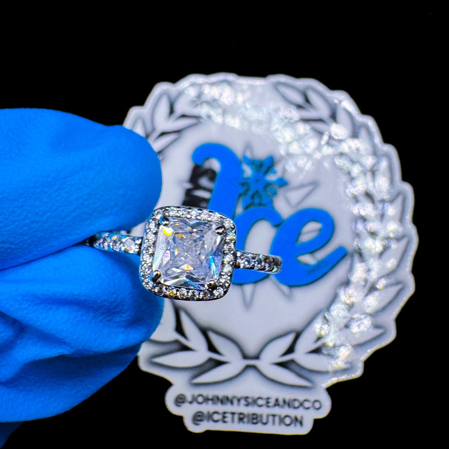 A two layer band ring with a large central diamond and smaller surrounding stones, displayed on a blue glove with a decorative background.