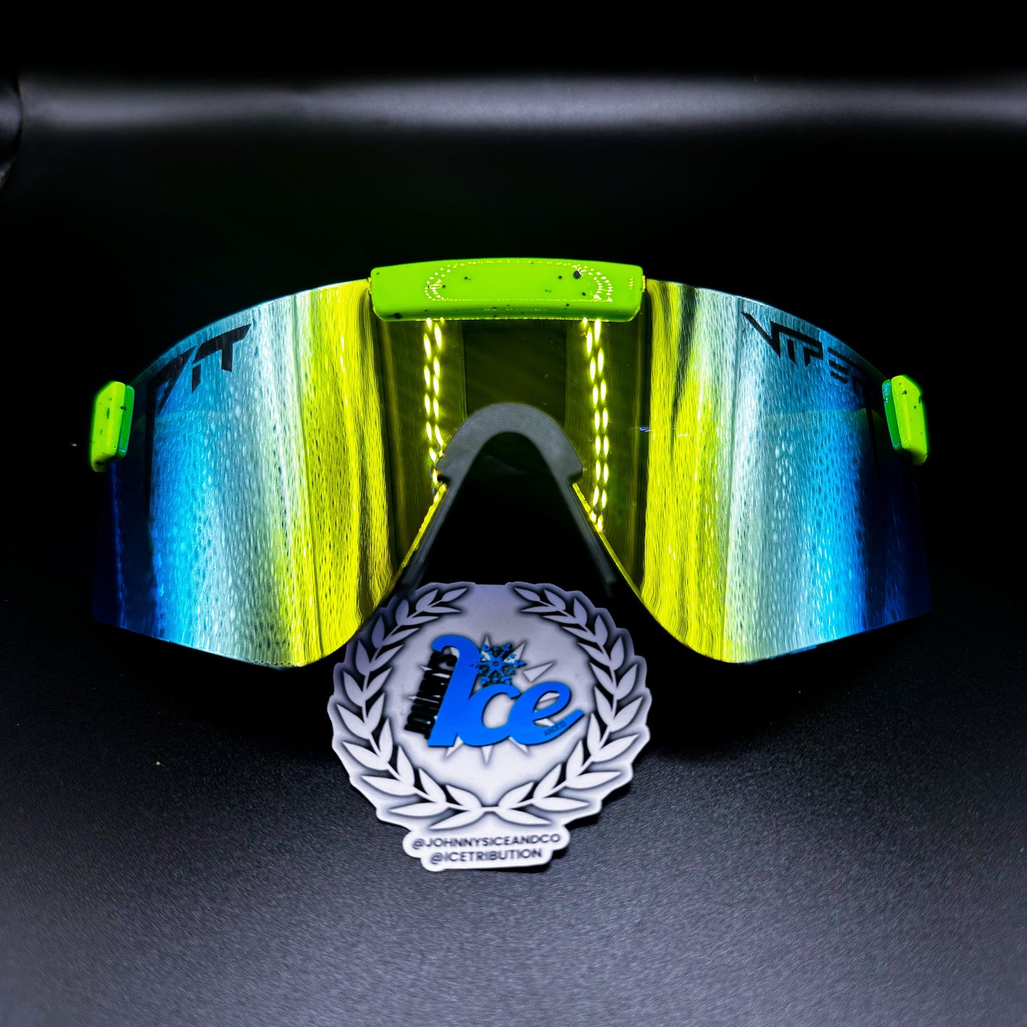 A pair of sunglasses with blue and green mirrored lenses, and yellow accents around the frame. The sunglasses are placed on a surface with a reflective badge in front displaying the brand 'Ice'.