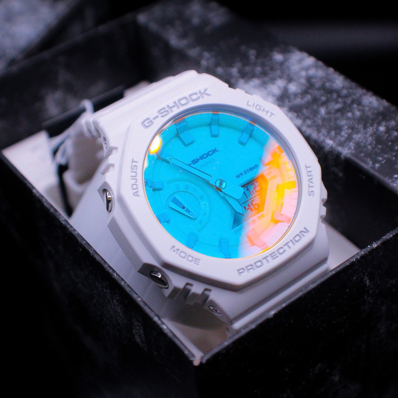 A G-Shock watch with a blue dial and white casing, illuminated by a blue light, displayed in an open box with black interior.