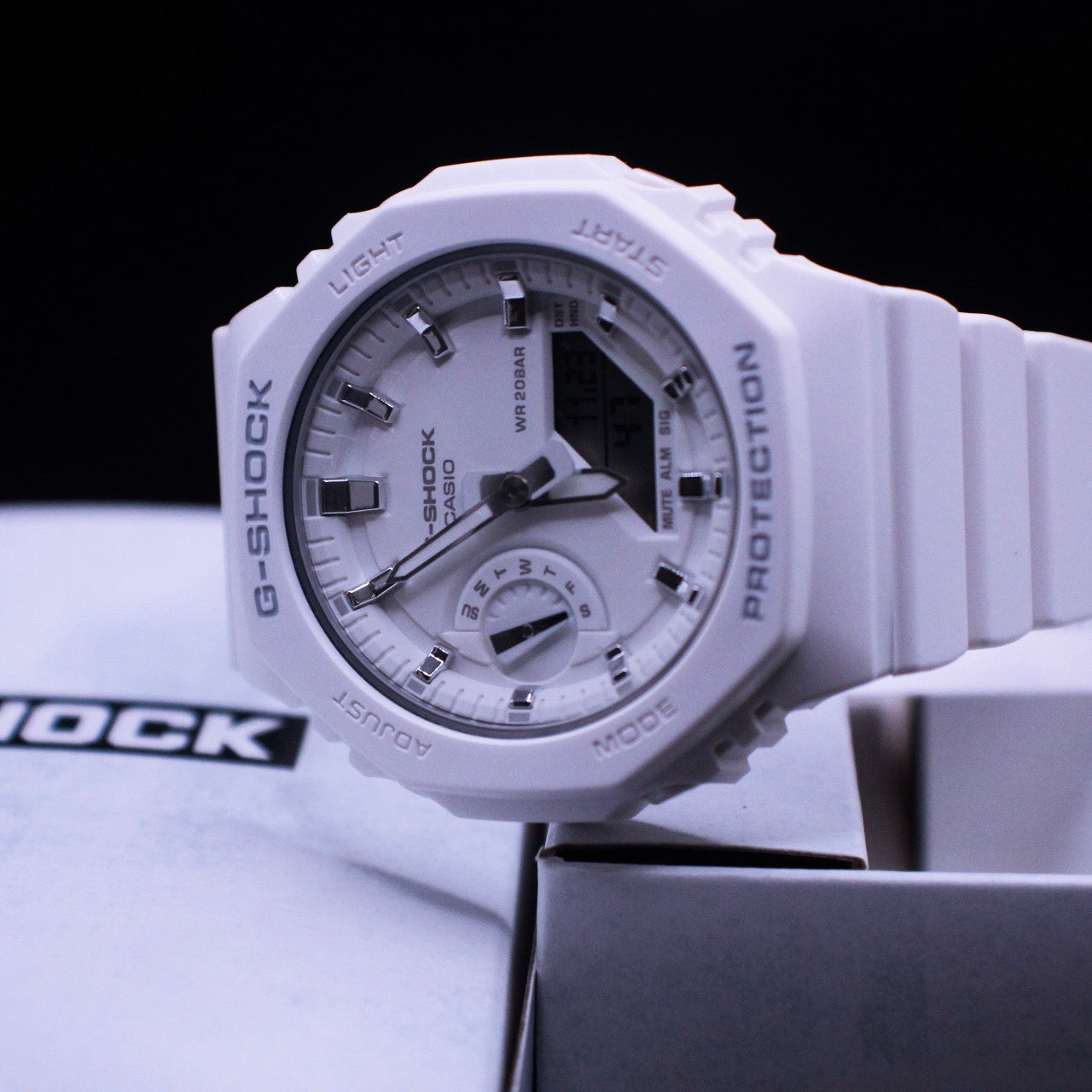 A white G-Shock watch with a baby blue illuminate band and white casing, placed on a box that has 'GSHOCK' written on it.
